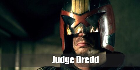Judge Dredd wears a dark helmet over his super hero outfit with padded vests, tops, and pants. He has a belt with green holsters and a gold badge.