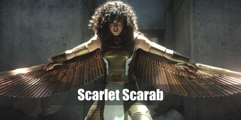Scarlet Scarab's costume features a metallic gold top with red details, gold tights, and boots. It is styled with gold accessories, belt, and wings, too. 