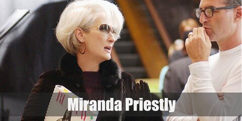 Miranda Priestly's outfit is a purple pencil dress with a black coat. She styles it with a pair of gloves, stockings, and heels. She also carries a white bag and has white hair.
