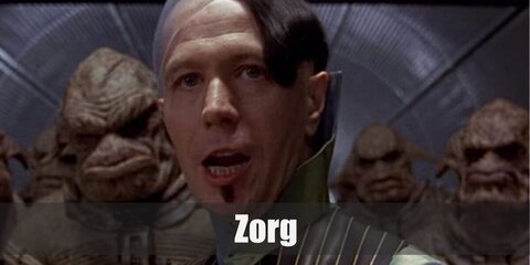 Jean-Baptiste Emanuel Zorg wears a green shirt under a striped vest with matching pants. He has a bald spot on his head and a mini moustache. He also carries a large gun.