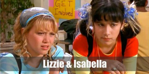 Lizzie McGuire’s costume is a silver cropped top, a silver jacket, silver pants, silver heels, and a blonde wig. Isabella’s costume is a green jacket, a green mini skirt, green sheer wrap-around, black fishnets, green knee-high boots, and a black wig.