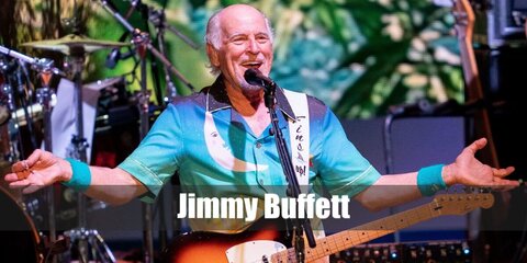 Jimmy Buffett wears a Hawaiian top with khaki shorts and he carries a guitar.
