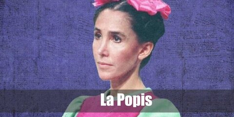 La Popis wears a pink dress with green sleeves. She wears white socks and a pair of dark shoes. La Popis also carries a doll.