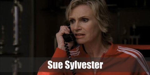 Sue Silvester wears a red tracksuit with white shoes and has short blonde hair.